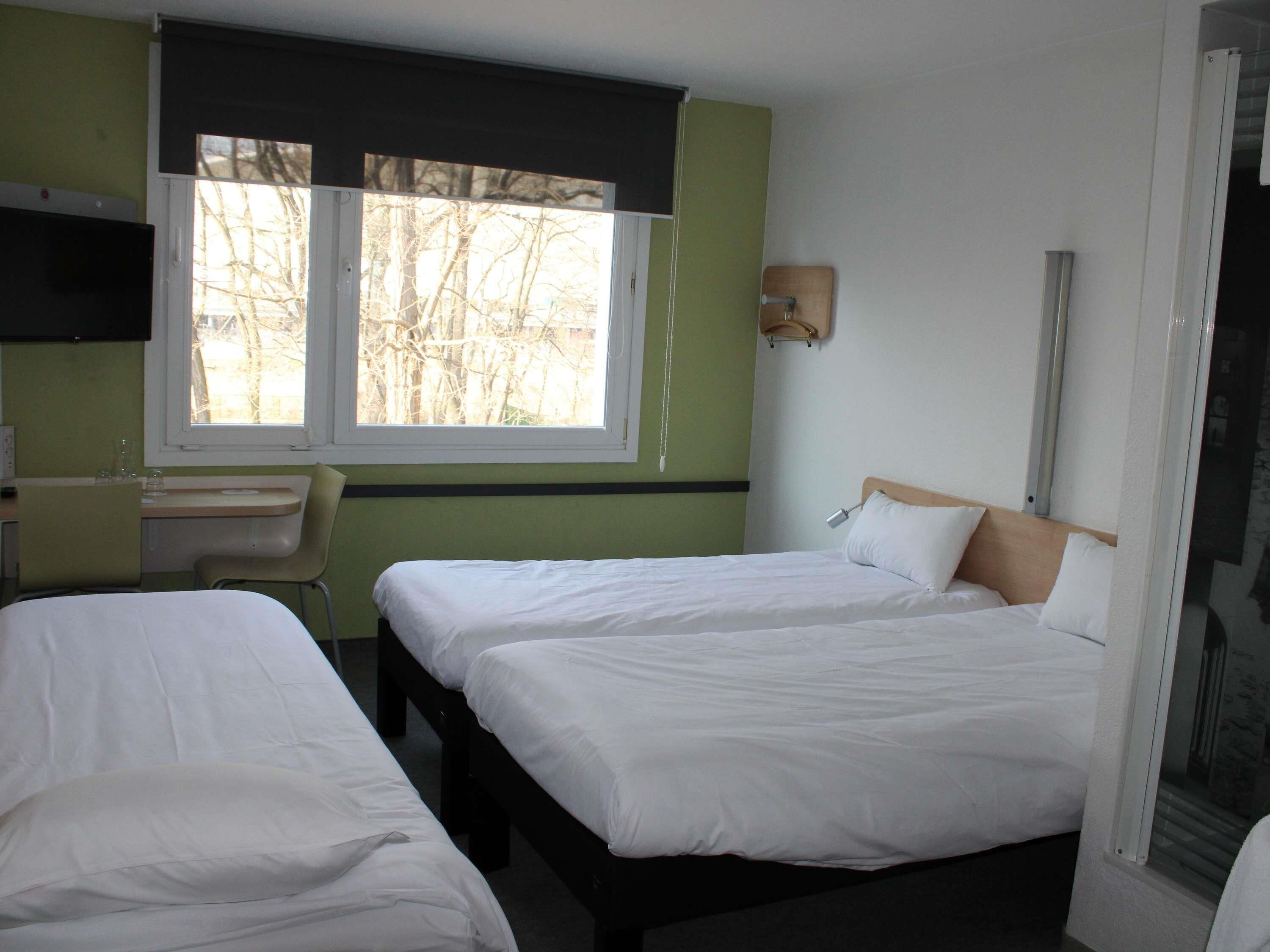 A hotel room with two beds, a desk, a chair, a window with a view of trees, and a wall-mounted lamp.