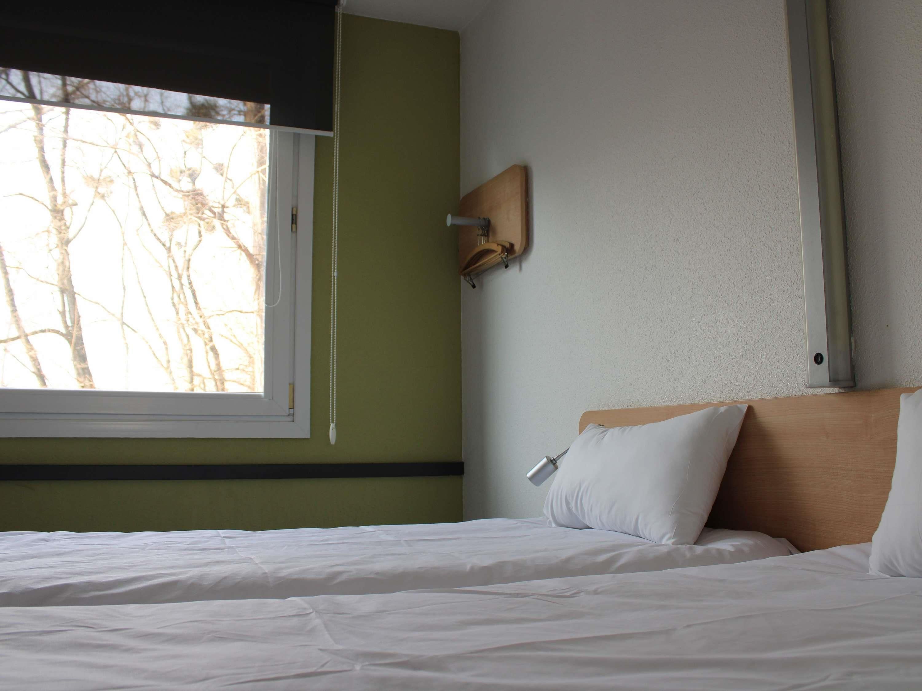 Et hotelværelse med seng, sengetæppe, lampe, vindue med udsigt til træer og vægmonteret hylde.
