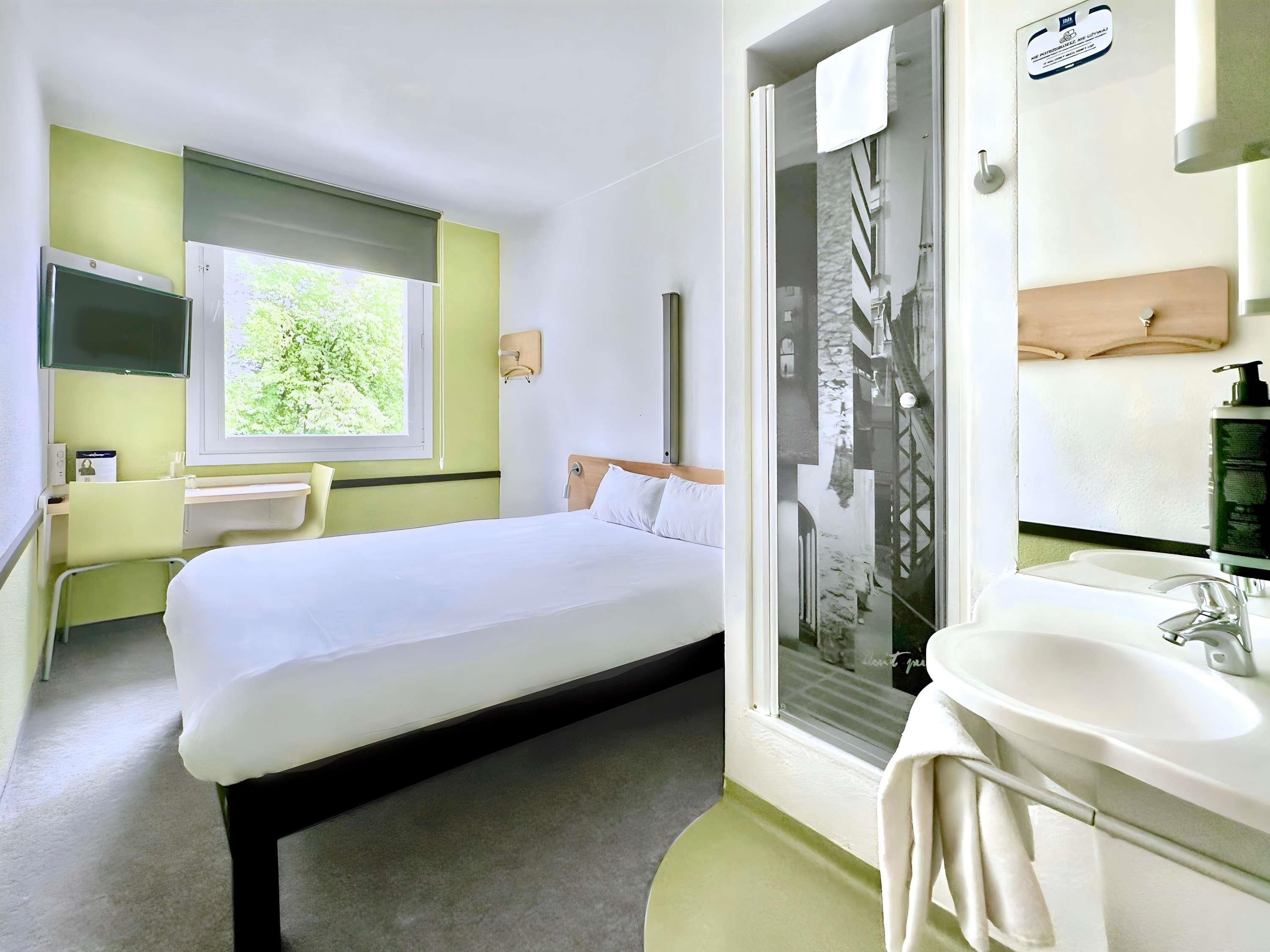 A hotel room with a bed, a window with a view of greenery, a shower area, and a bathroom with a sink and mirror.