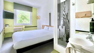 A hotel room with a bed, a window with a view of greenery, a shower area, and a bathroom with a sink and mirror.