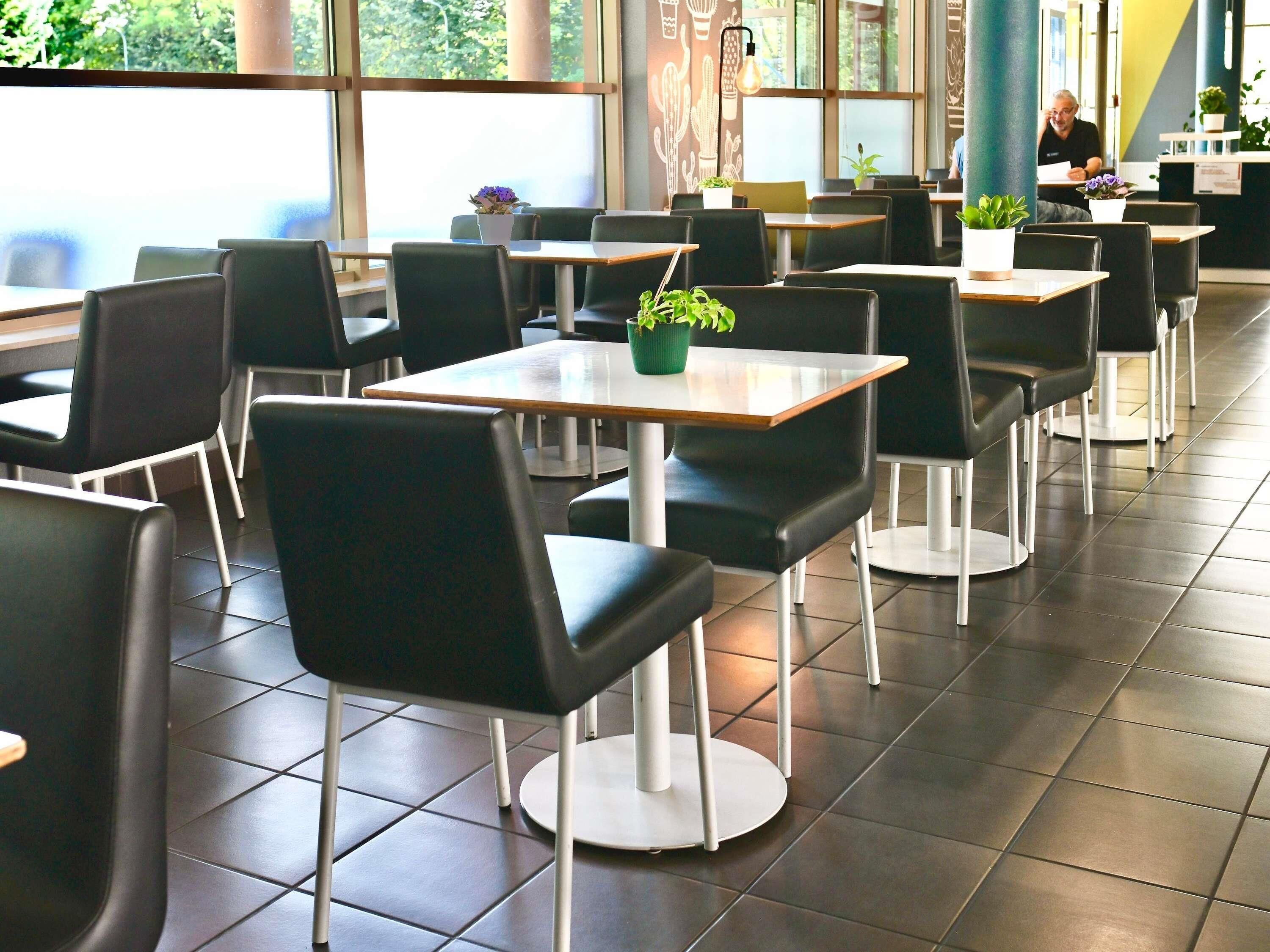 A spacious indoor seating area with multiple tables and chairs, large windows, and potted plants.