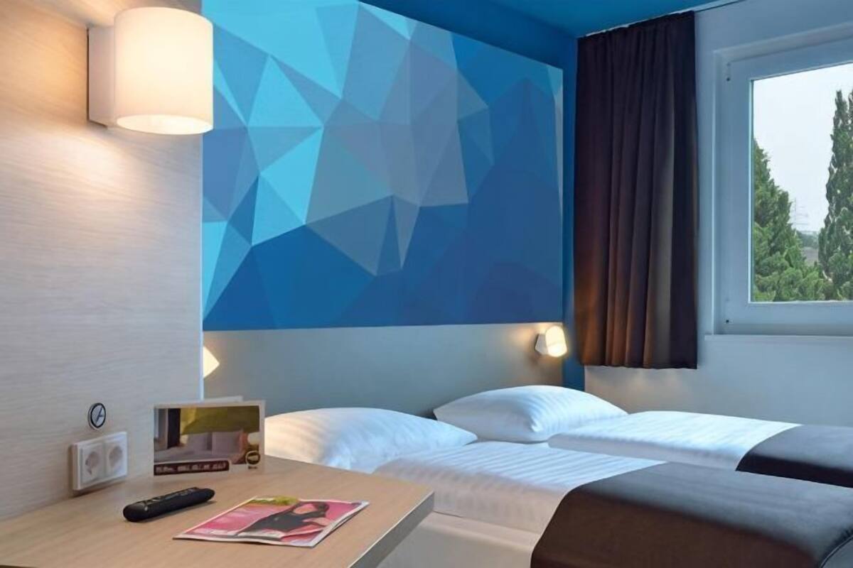 Una habitación de hotel con cama, mesita de noche, lámpara y ventana con cortinas.