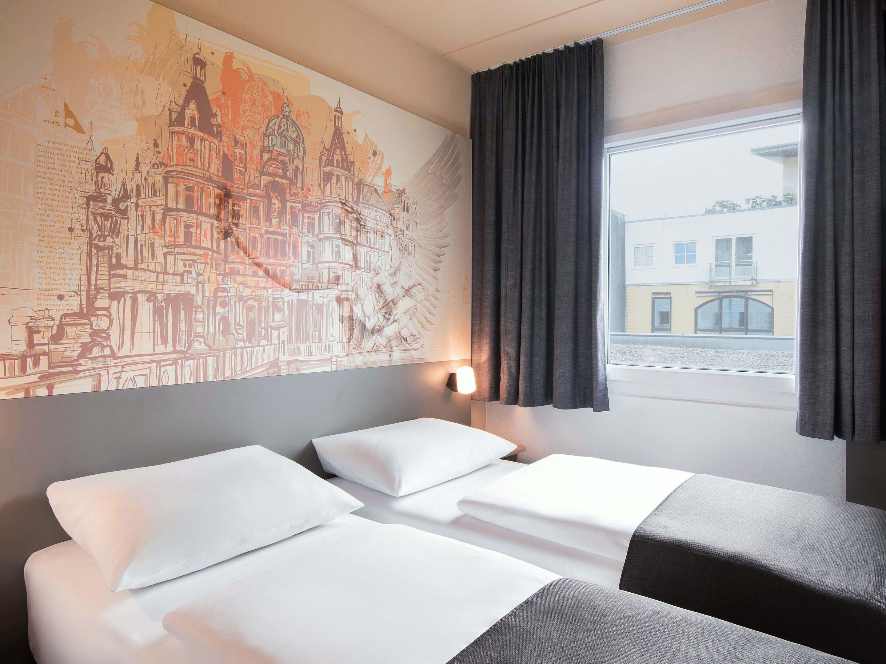 Une chambre d’hôtel avec deux lits, une grande fresque représentant un paysage urbain sur le mur, une fenêtre avec des rideaux et une vue sur les bâtiments à l’extérieur.