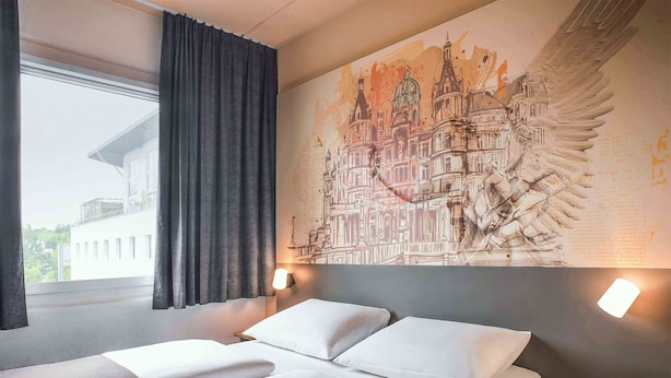 A hotel room with a bed, a large wall mural, and a window with curtains. - B&B HOTEL Schwerin-Süd (Schwerin)