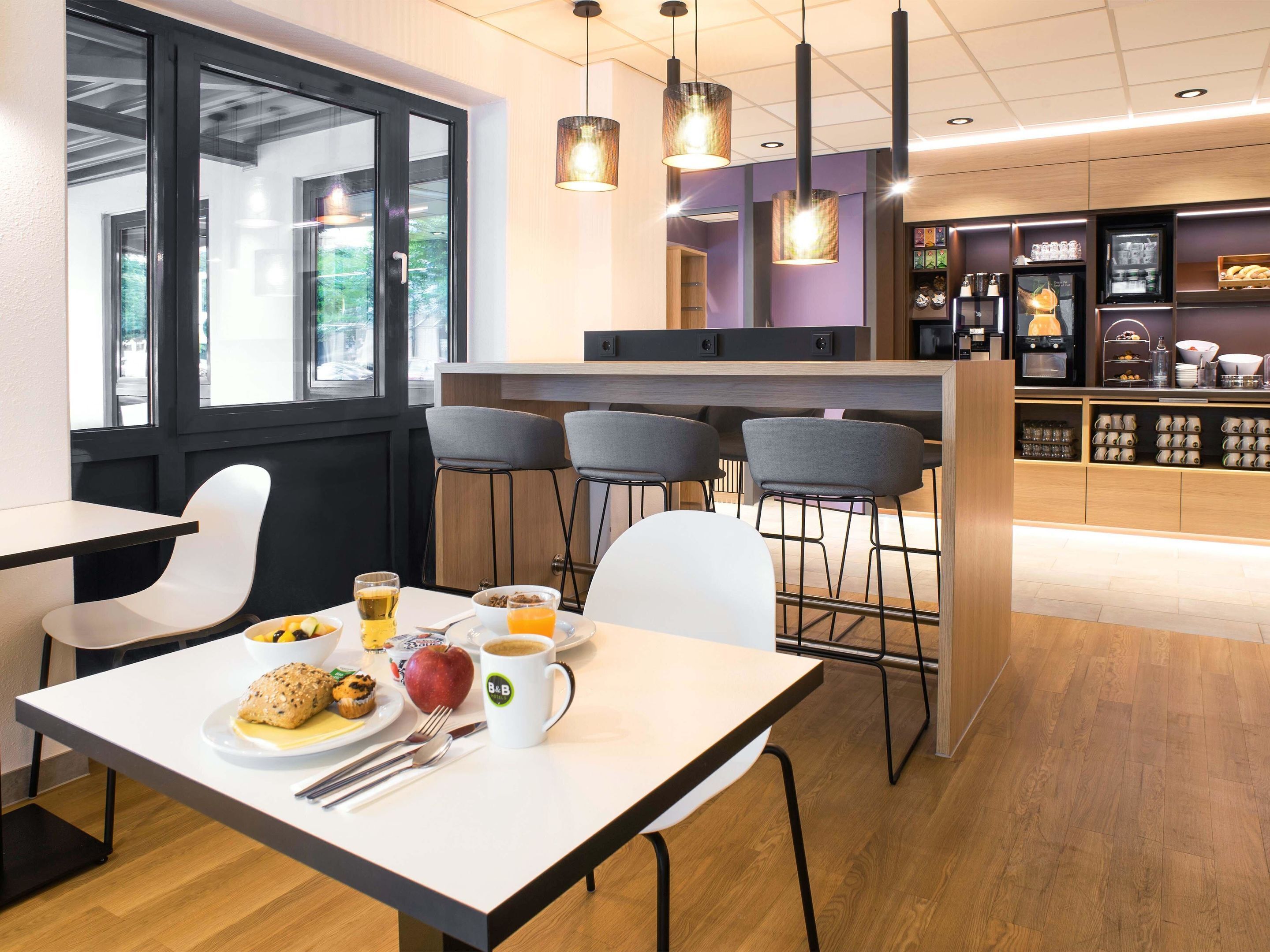 Un café moderne avec une table blanche, des chaises et une table dressée pour le petit-déjeuner, comprenant une assiette de nourriture, une tasse et un verre de jus d’orange.