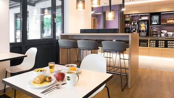 Un café moderne avec une table blanche, des chaises et une table dressée pour le petit-déjeuner, comprenant une assiette de nourriture, une tasse et un verre de jus d’orange.