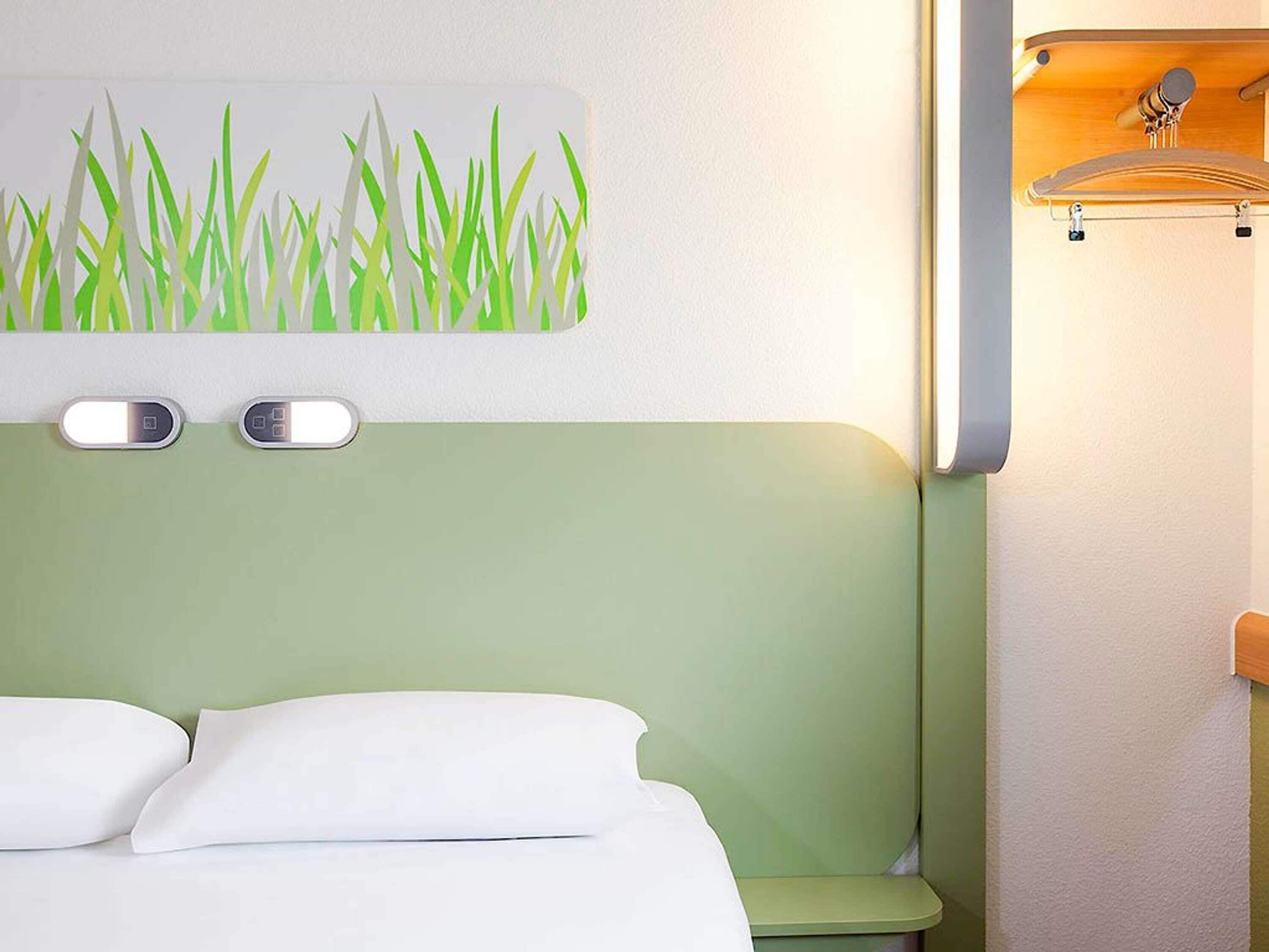 Une chambre d’hôtel avec un lit, une tête de lit dotée d’un panneau vert clair, une œuvre d’art représentant de l’herbe accrochée au mur, et une petite étagère avec un crochet.