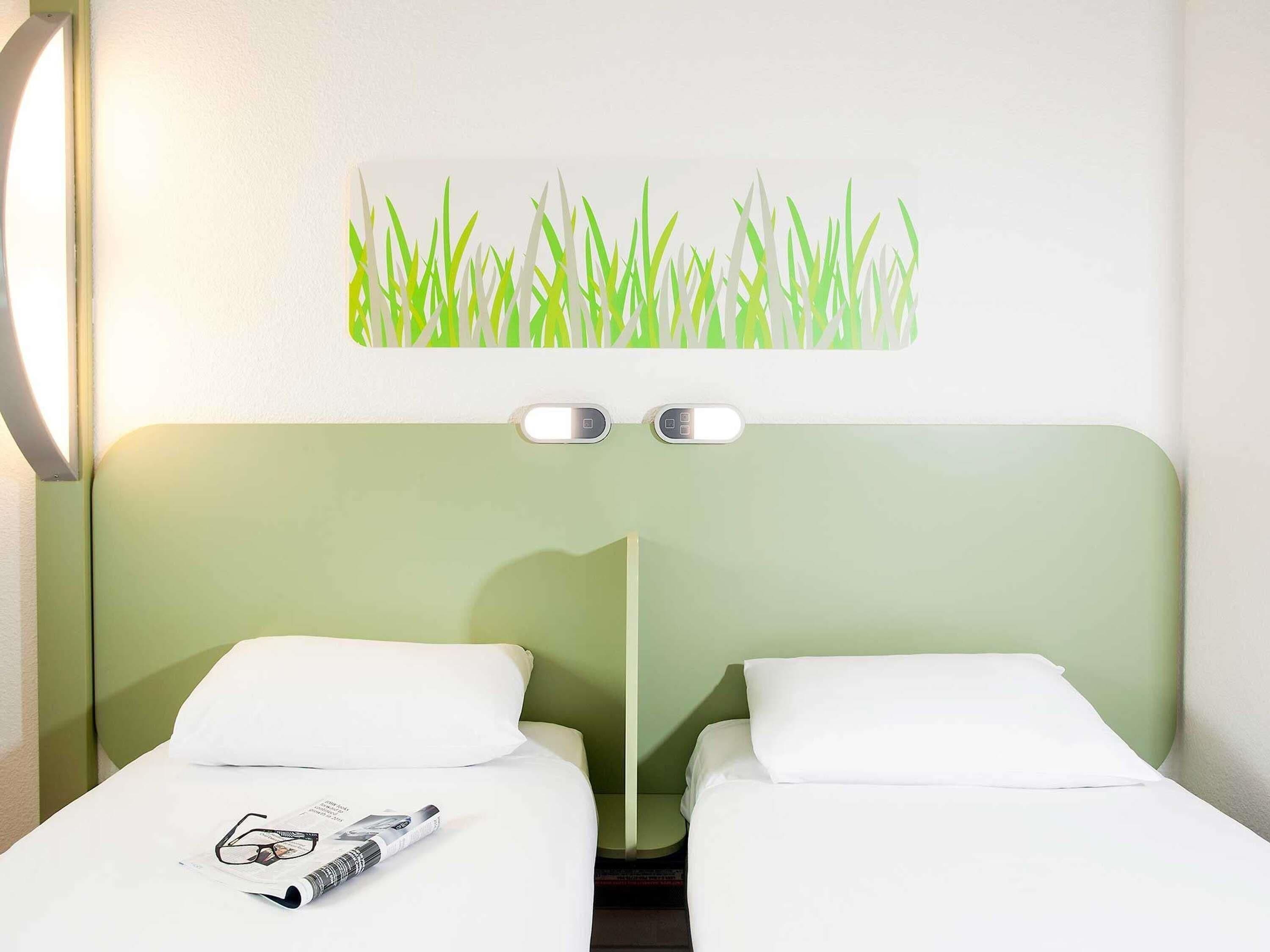 Une chambre d’hôtel avec deux lits simples, une tête de lit verte, une lampe fixée au mur et un tableau représentant un champ d’herbe.
