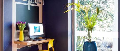 Un espacio de trabajo con una computadora, una silla amarilla, un florero con flores moradas y un ventanal con vistas a árboles.