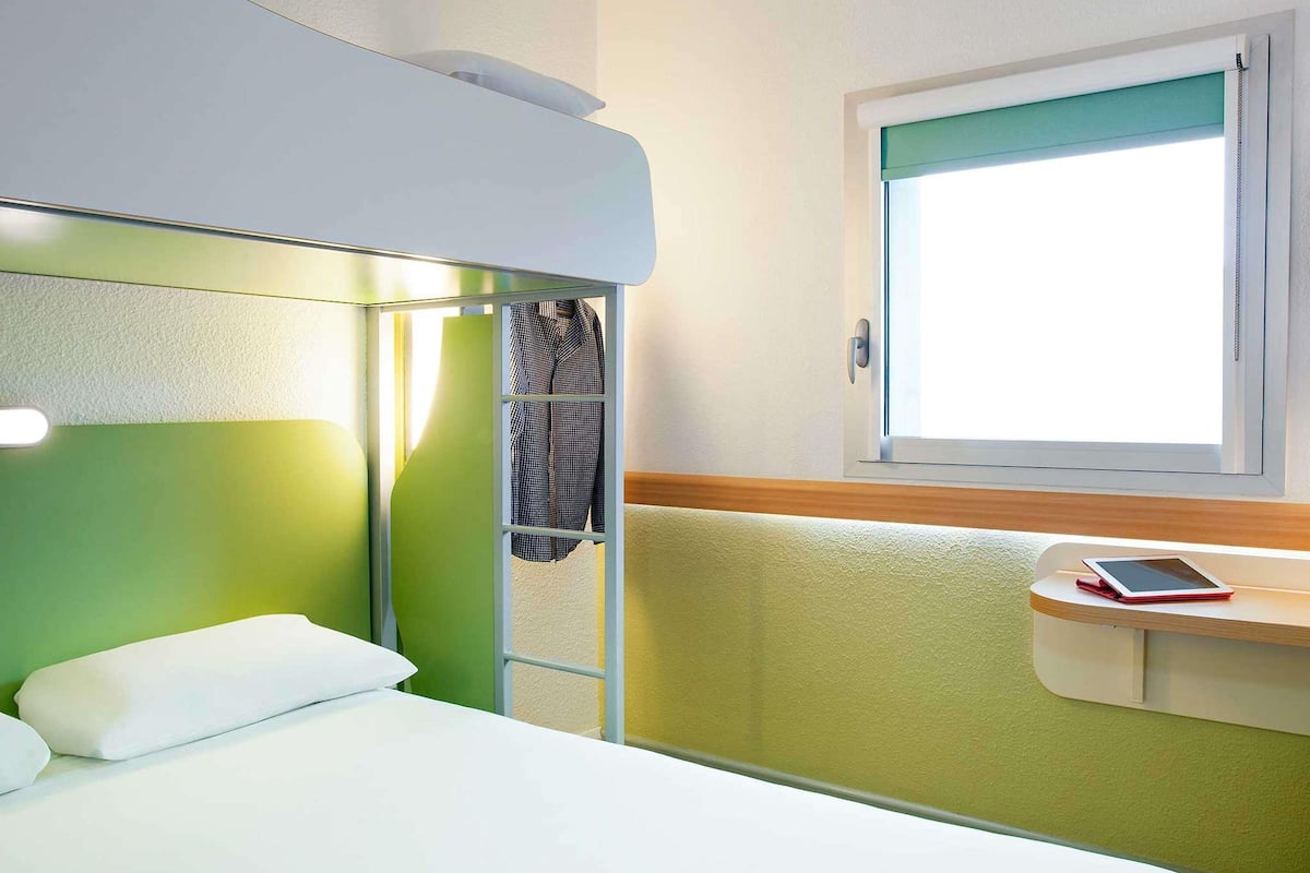 Habitación de hotel con literas, una mesita de noche con una tableta, una ventana con vistas y una pared verde.