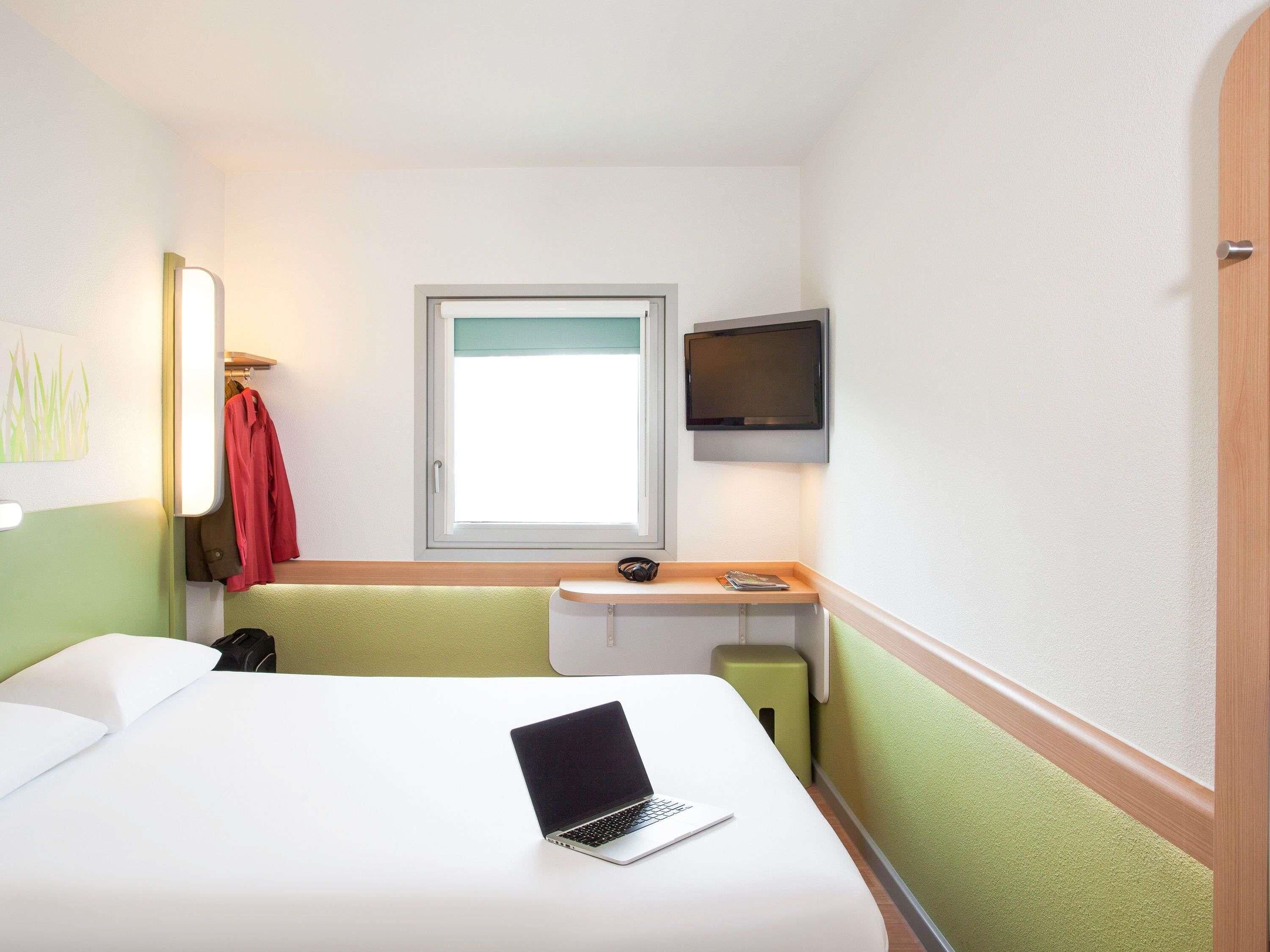 Una habitación de hotel con una cama, un escritorio con una computadora portátil, un televisor y una ventana.