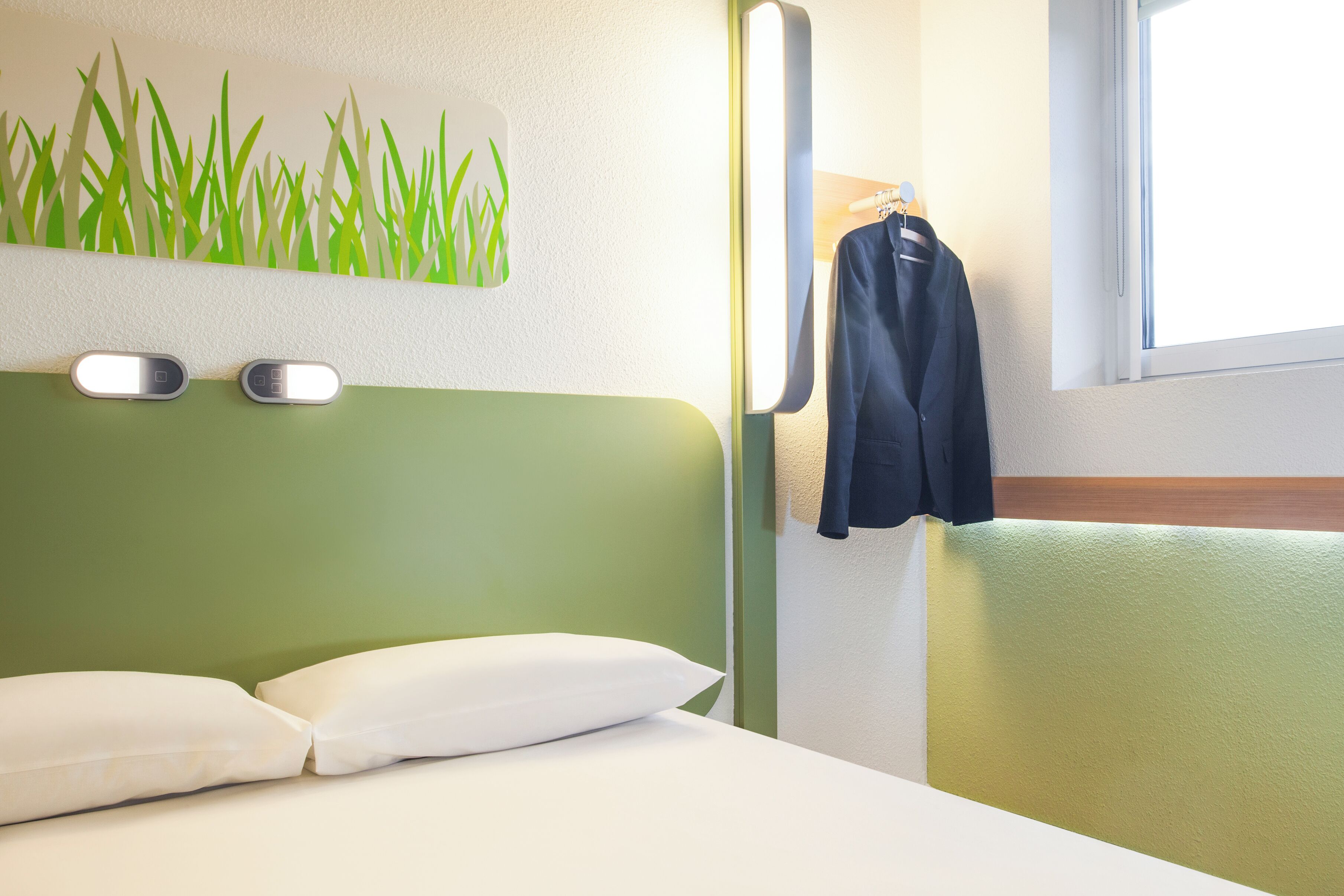 Habitación de hotel con cabecera verde, almohadas blancas, una lámpara de pared y ventana con vistas.