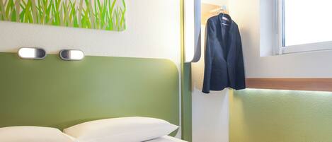 Habitación de hotel con cabecera verde, almohadas blancas, una lámpara de pared y ventana con vistas.