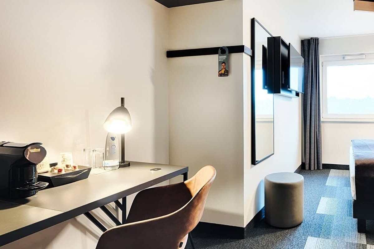 A hotel room with a bed, a desk with a coffee maker, a chair, a round ottoman, a TV mounted on the wall, and a window with curtains.