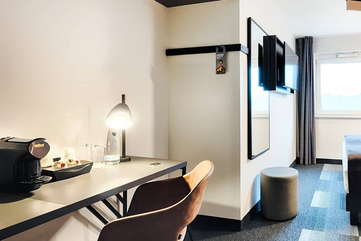 A hotel room with a bed, a desk with a coffee maker, a chair, a round ottoman, a TV mounted on the wall, and a window with curtains.