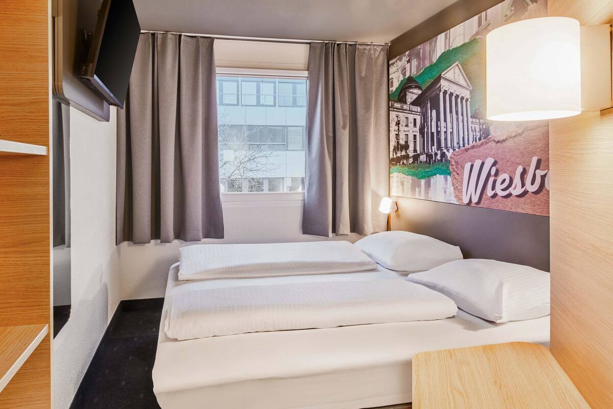 Une chambre d’hôtel avec un lit, des rideaux, une fenêtre, une lampe fixée au mur et une pancarte portant le mot « Wiesbaden ».