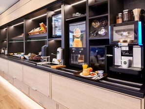 A modern breakfast bar with a coffee machine, a variety of food items, and a fruit display. - B&B HOTEL Wiesbaden-Ost (Wiesbaden)