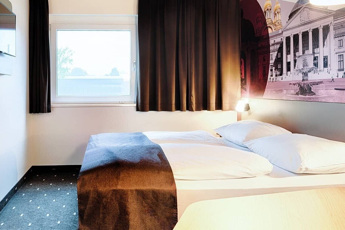 A hotel room with a bed, a TV, a window with curtains, a wooden shelf, and a wall-mounted picture.