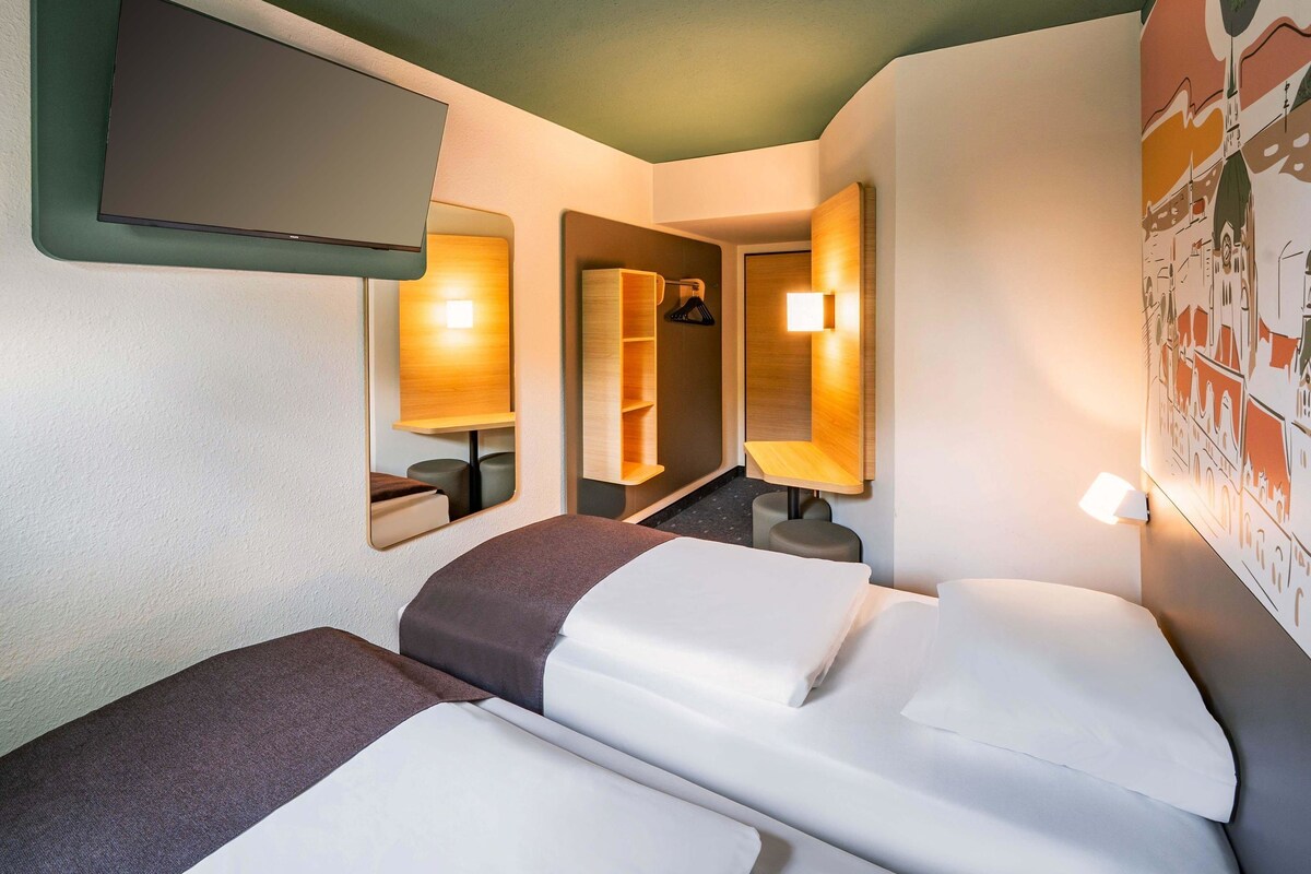 A hotel room with two beds, a flat-screen TV, wall-mounted lamps, and a cityscape mural.