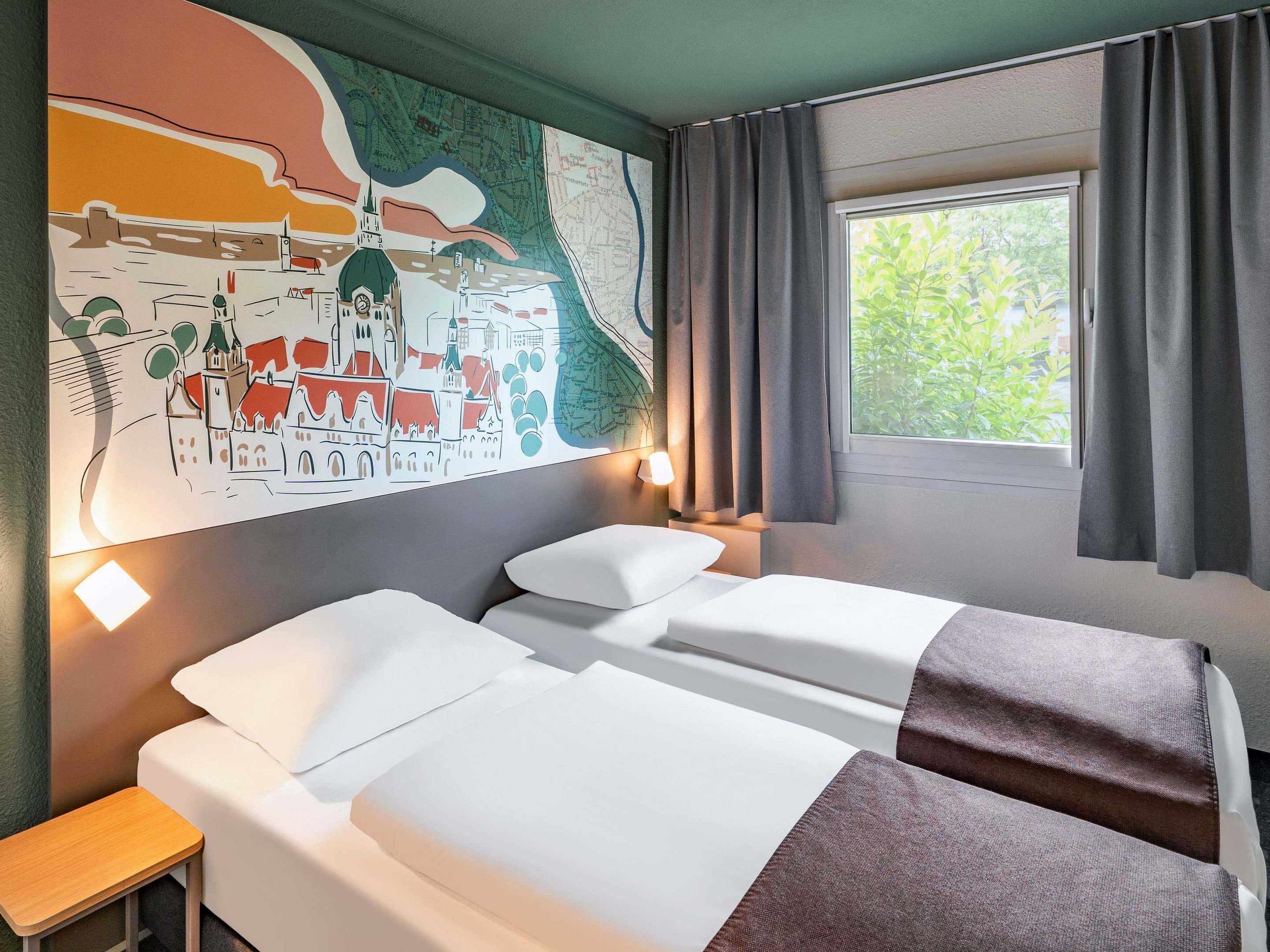 A hotel room with two beds, a map artwork on the wall, a window with curtains, and a bedside table with a lamp.