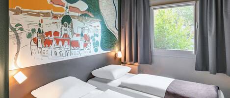 A hotel room with two beds, a map artwork on the wall, a window with curtains, and a bedside table with a lamp.