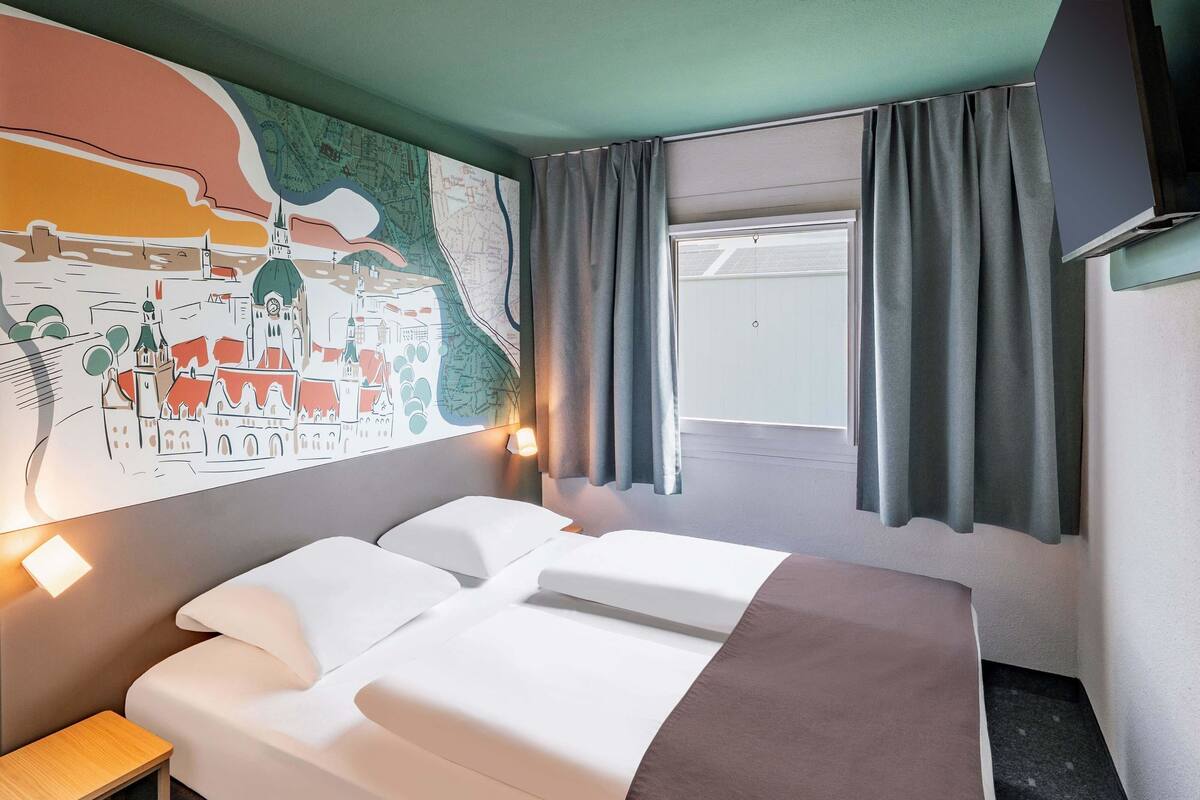 Habitación de hotel con una cama grande, una mesita pequeña, un mapa en la pared y una ventana con cortinas.