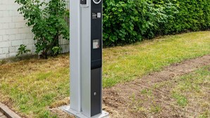 Una estructura metálica alta y rectangular con un código QR y un elemento circular, probablemente un quiosco de servicios públicos o información, frente a un seto y un muro de ladrillo.