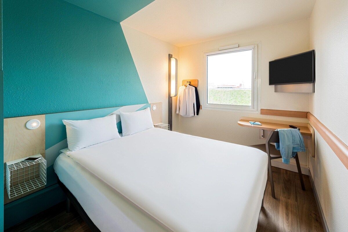 Ein Hotelzimmer mit Bett, Nachttisch, an der Wand montiertem Fernseher, Fenster und kleinem Schreibtisch mit Stuhl.