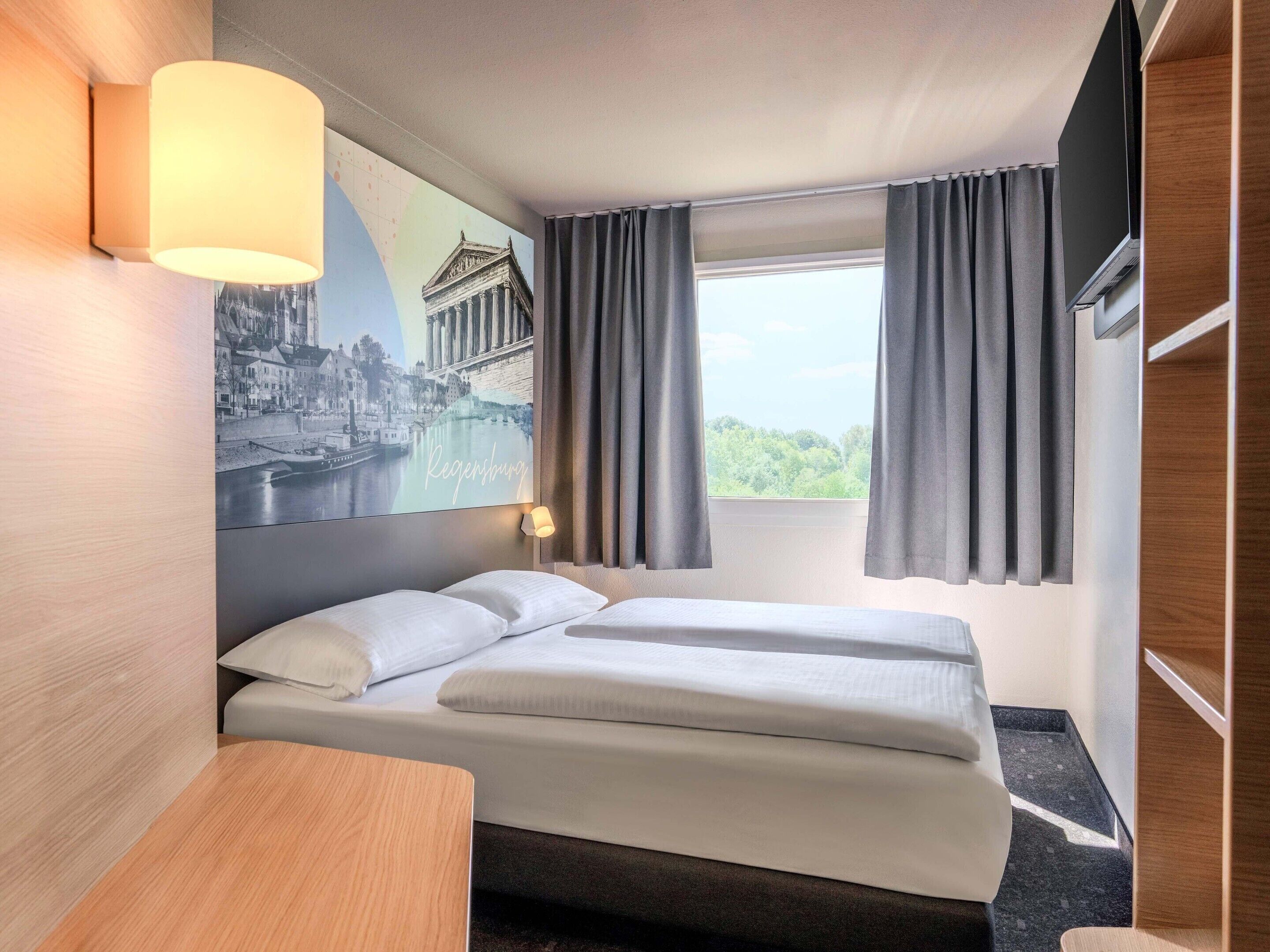 Ein Hotelzimmer mit einem Bett, einem Schreibtisch, einer Lampe, einem fensterbelaubten Fenster und einem an der Wand montierten Fernseher.