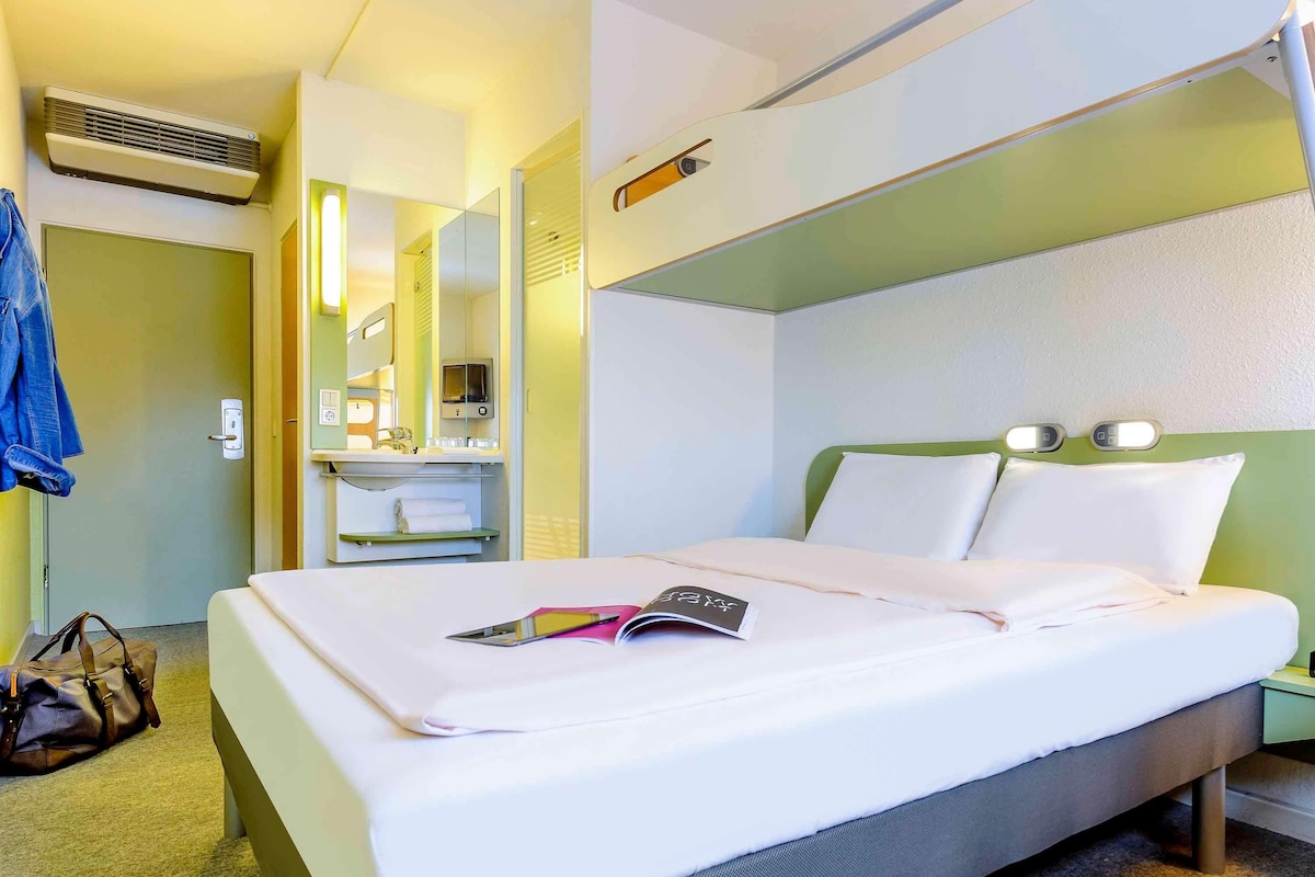 Une chambre d’hôtel comprenant un lit, une table de chevet, un miroir, une salle de bain avec un lavabo et un appareil de climatisation.