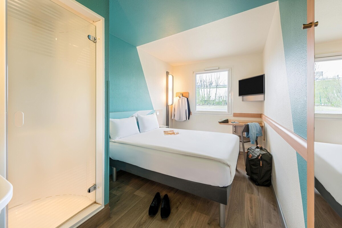 Une chambre d’hôtel de taille réduite, comprenant un lit, un petit bureau, une chaise, un téléviseur et une fenêtre donnant sur la verdure.