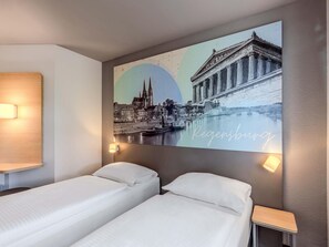 A hotel room with two beds, a wall-mounted artwork of a cityscape, and a small bedside table. - B&B HOTEL Regensburg-Ost (Regensburg)