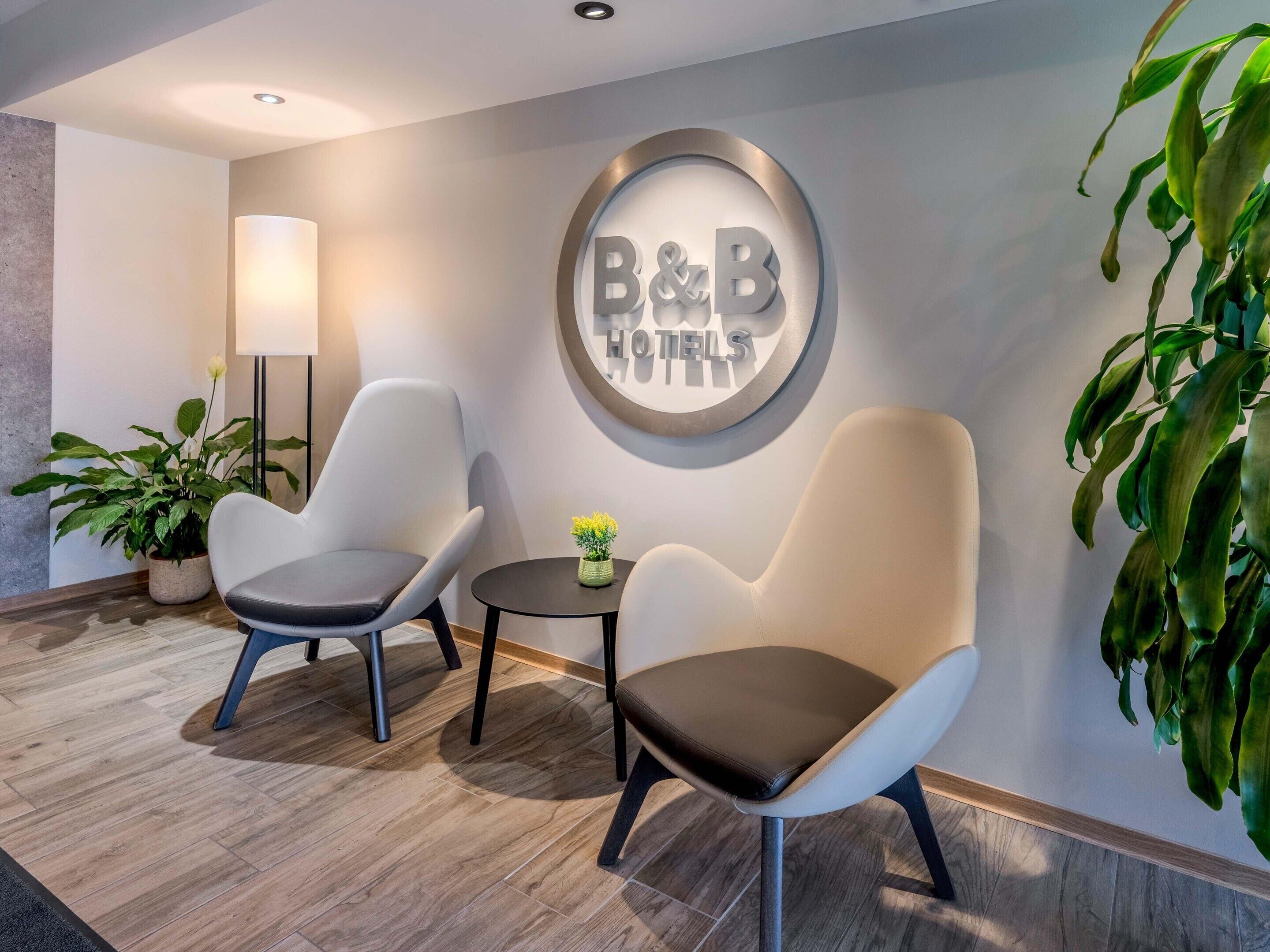 A hotel lobby with two armchairs, a round wall sign reading 'B&B HOTELS', a floor lamp, and a potted plant.