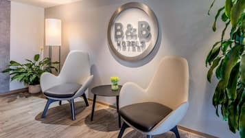 A hotel lobby with two armchairs, a round wall sign reading 'B&B HOTELS', a floor lamp, and a potted plant.