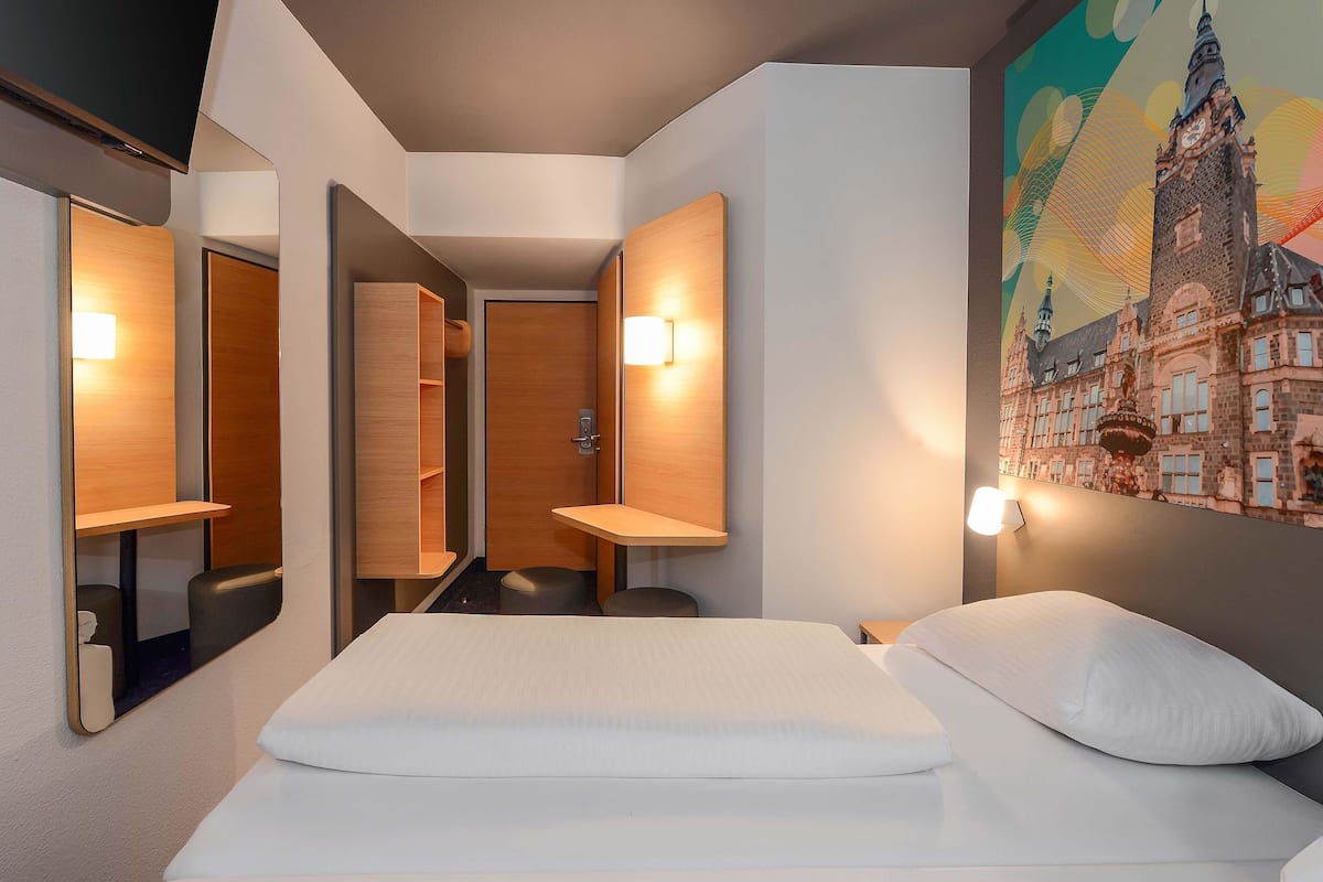 A hotel room with a bed, a mirror, a wooden cabinet, and a wall-mounted light fixture.