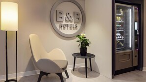 Eine Hotelrezeption mit einem modernen Sessel, einer Topfpflanze, einem Schild mit der Aufschrift "B&B HOTELS" und einem Verkaufsautomaten.