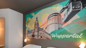 Ein Hotelzimmer mit einem Bett, einer Wandleuchte und einem großen Wandbild eines Gebäudes mit dem Text 'Wuppertal'.