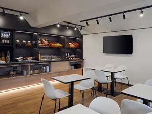 A modern dining area with white chairs and tables, a buffet with various food items, and a flat-screen TV on the wall. - B&B HOTEL Wuppertal-Oberbarmen (Wuppertal)
