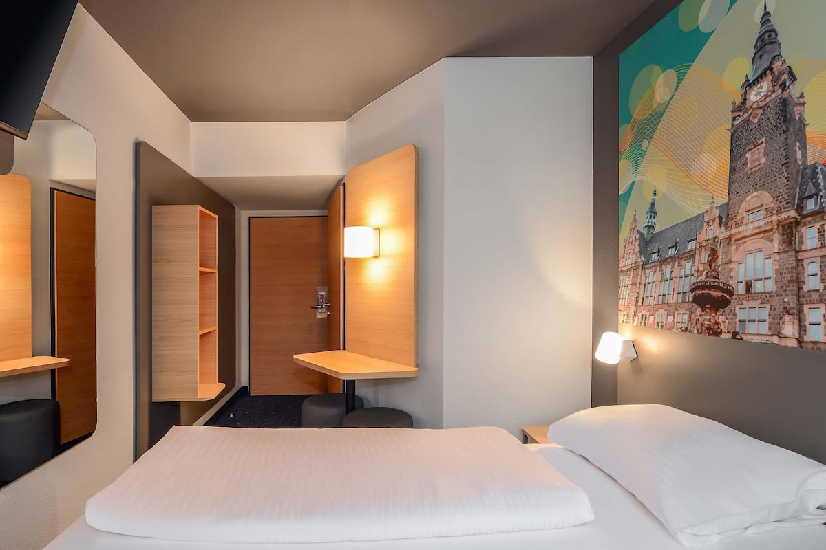 A hotel room with a bed, a wooden wardrobe, a mirror, a wall-mounted light, and a large painting of a historic building.