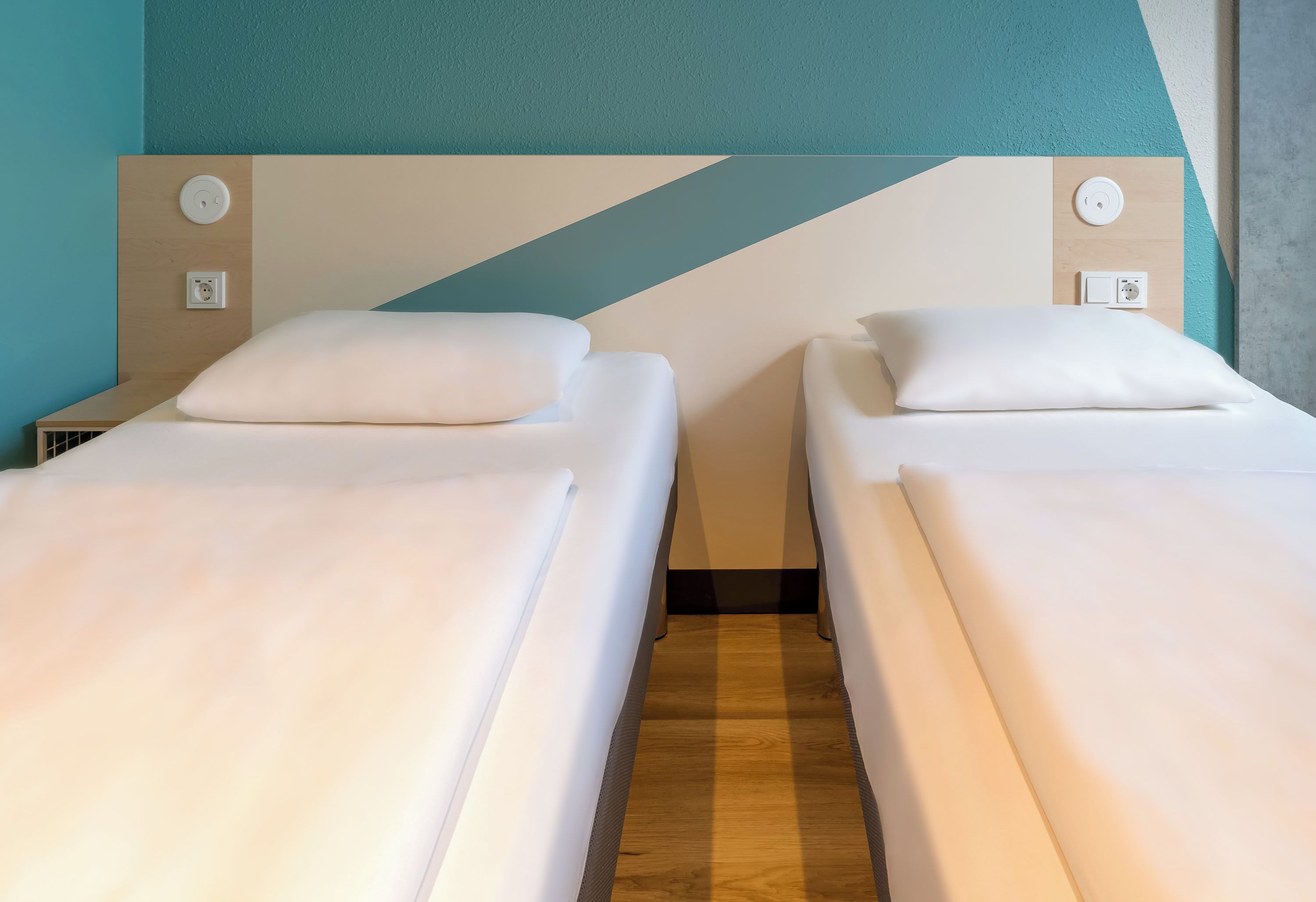 A hotel room with two single beds, a wooden floor, and a teal accent wall.