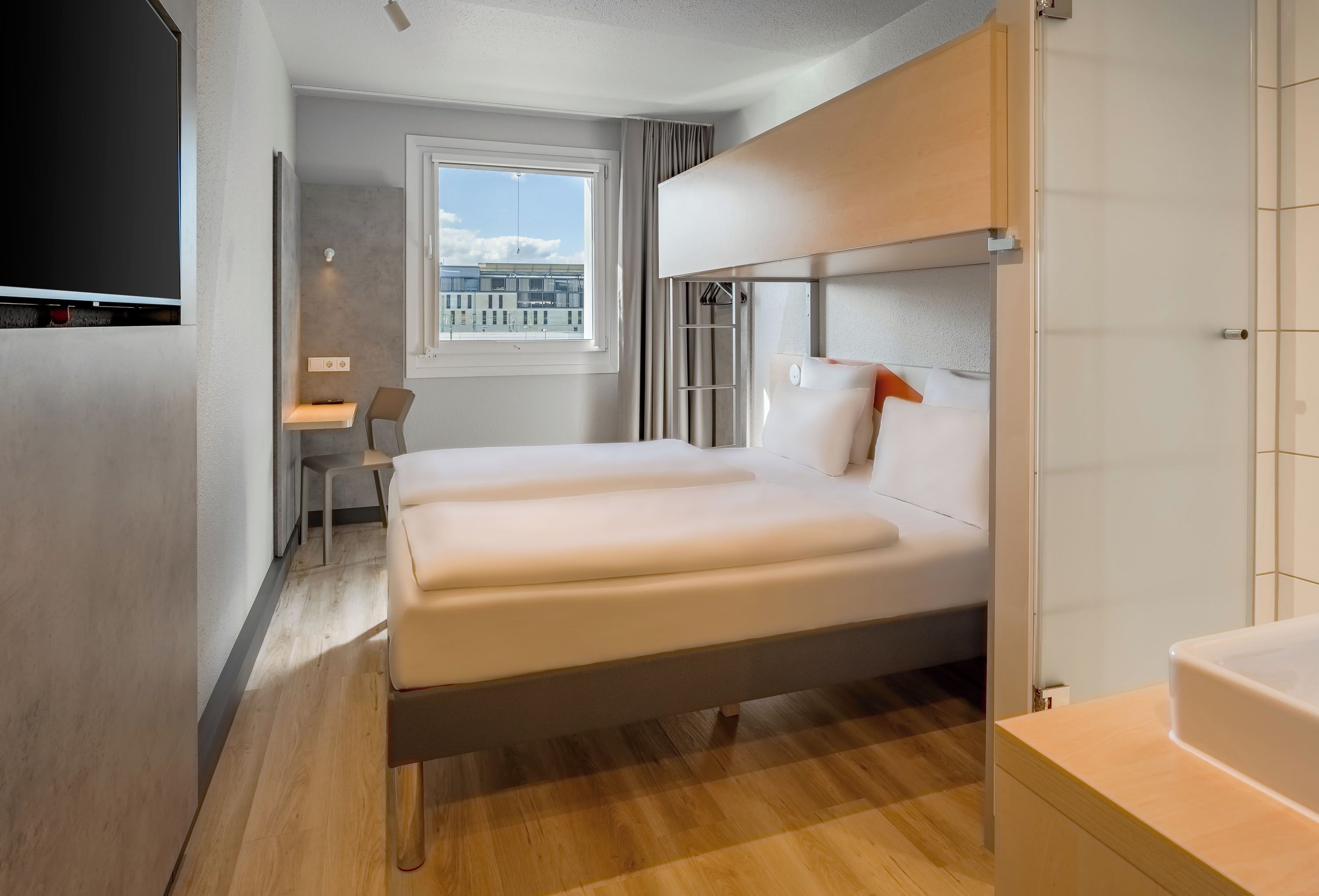 Una habitación de hotel moderna con una cama grande, un escritorio y un ventanal con vistas a edificios.