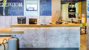 A reception desk with a computer, a television, and a seating area with chairs. - Ibis Budget Hannover Hauptbahnhof (Hannover)