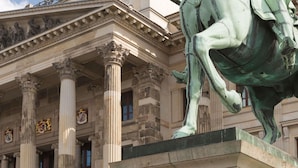 Fachada clássica de edifício com colunas, uma estátua de cavalo e uma placa com a inscrição 'HERZOG FRIEDRICH WILHELM'.