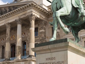 Eine klassizistische Gebäudefassade mit Säulen, einer Pferdestatue und einer Tafel mit der Aufschrift 'HERZOG FRIEDRICH WILHELM'.