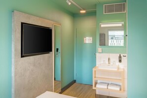 Un baño con un televisor montado en la pared, un lavamanos con un espejo encima y una puerta que da a otra habitación.