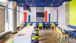 A modern dining area with a long table, colorful chairs, and a large window. - Ibis Budget Hannover Hauptbahnhof (Hannover)