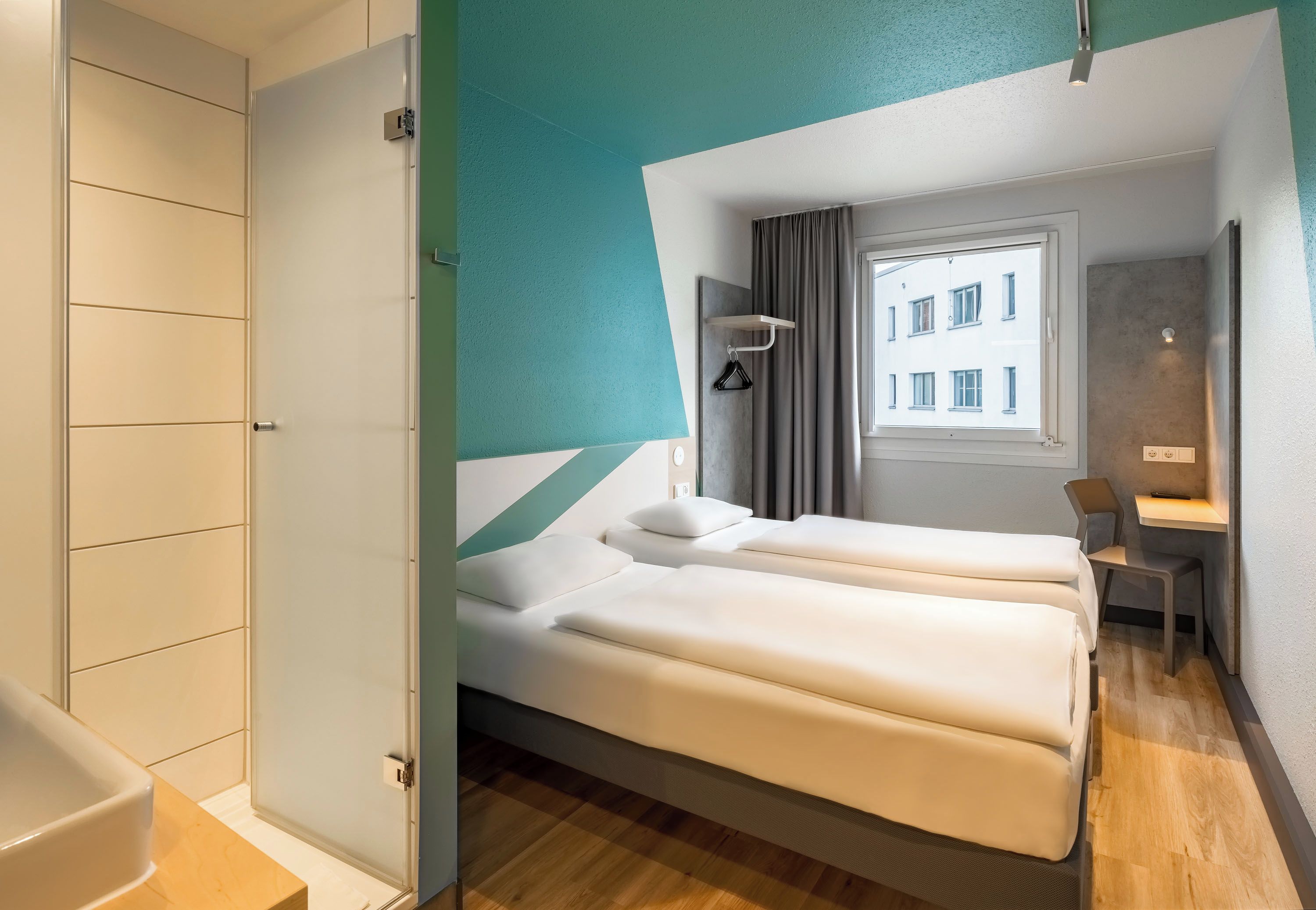 A hotel room with a bed, a desk, and a view of a building through a window.