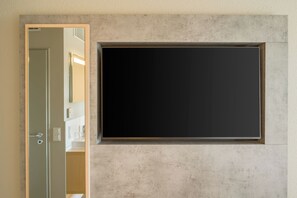 A room with a large, dark-colored flat-screen TV mounted on a textured wall, a door leading to another room, and a small shelf with a few items. - Ibis Express Hannover Central Station (Hannover)