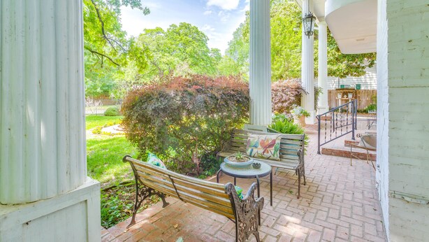 A covered patio with a bench and table, surrounded by greenery and a white building. - Azalea Plantation B&B (Fort Worth)