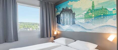 A hotel room with a bed, a window with curtains, a wall-mounted lamp, and a large mosaic artwork.